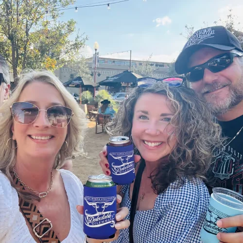 Three smiling guests taking a selfie on an outdoor patio, holding Delectable Dilemmas koozies