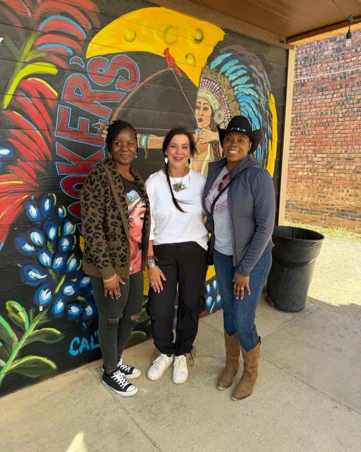Three women smiling in front of a colorful painted mural in the Stockyards