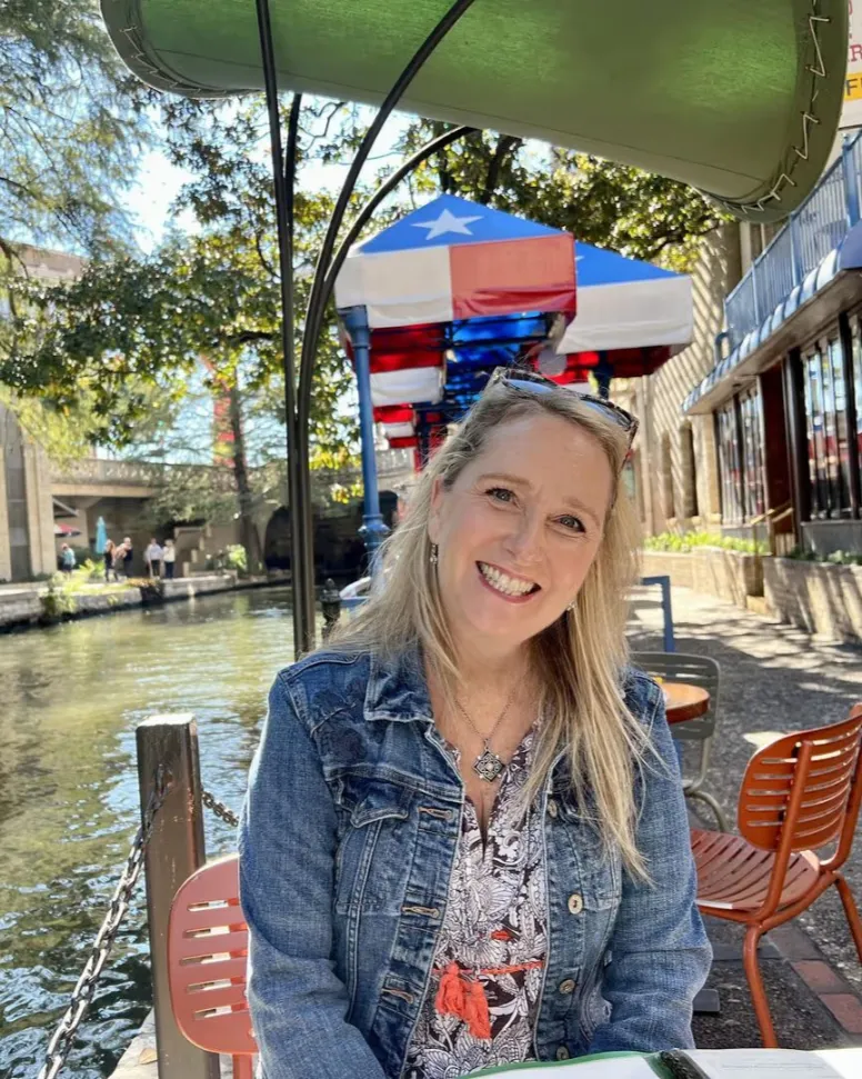 A smiling woman in a denim jacket sitting at a canal-side table with outdoor chairs behind her