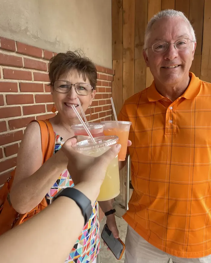 A couple smiling as they toast with three colorful iced drinks in front of a brick wall