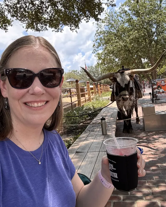 A smiling guest in sunglasses holding a cold drink with a Texas longhorn standing behind her on a Stockyards street